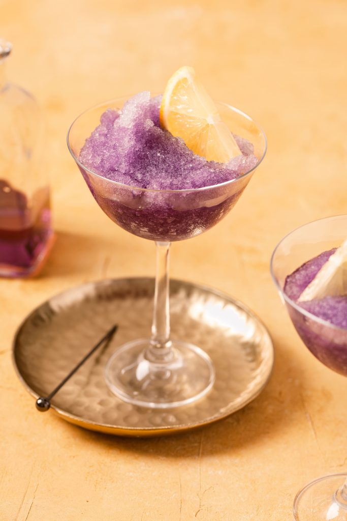 Two stemmed glasses with purple slush and lemon slices on a yellow background.