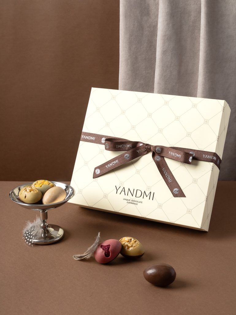 Предметна зйомка подарункової коробки шоколадних цукерок Yandmi з декоративною стрічкою та кольоровими драже у рекламній фуд-зйомці для шоколадного бренду.